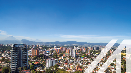Stadtansicht von Guatemala City: Hochhäuser umgeben von niedrigen Häusern, durchbrochen von grünen Bäumen, im Hintergrund Bergkette unter blauem Himmel