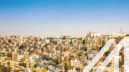 Stadtansicht von Amman: Blick auf viele helle Häuser unter blauem Himmel, eine Straße schlängelt sich bergauf. Über das Bild wurde ein weißes Austria A gelegt.