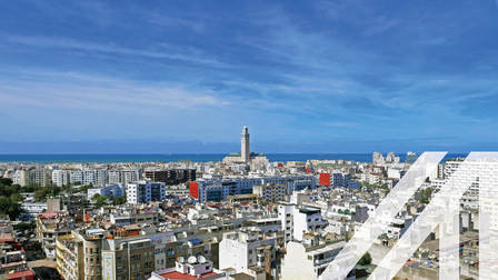 Panoramablick auf Casablanca, der Turm der Hassan II Moschee ragt hervor, unter blauem Himmel am Meer gelegen.
