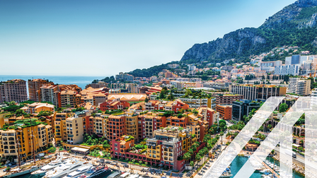 Blick auf den Hafen von Monte Carlo mit bunten Hochhäusern und Yachten, im Hintergrund sieht man das offene Meer und eine ansteigende bewaldete Bergkette  