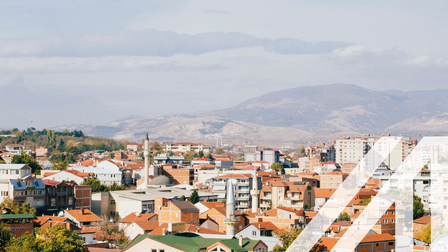 Stadtansicht von Skopje: weiße Häuser, rote Dächer, dazwischen Türme einer Moschee und Hochhäuser vor einer Bergkette unter grauem Himmel. Über das Bild wurde ein weißes Austria A gelegt.