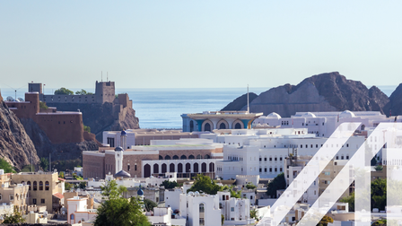 Historische Aldstadt von Maskat mit weißen orientalischen Häusern und einer historischen Festung links im Bild. Im Hintergrund sieht man den Golf von Oman und einen Berg. 

