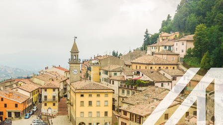 Stadtansicht von San Marino: Vorwiegend gelbbeige Häuser auf Hügel verlaufend mit Schindeldächern, Uhrturm hervorragend, im Zentrum Platz mit parkenden Autos, daneben Straße und grüner Laubbaumwald
