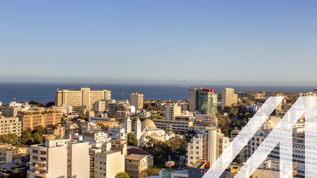 Stadtansicht von Dakar: moderne überwiegend helle Hochhäuser am Meer gelegen unter blauen Himmel. Über das Bild wurde ein weißes Austria A gelegt.