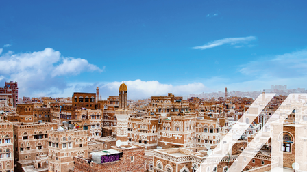 Stadtansicht von Sanaa: Die Straßen der Altstadt von Sanaa, der Hauptstadt des Jemen. Terrakottafarbene Häuser mit weißen Verzierungen unter blauem Himmel, in der Mitte ein Turm. Über das Bild wurde ein weißes Austria A gelegt.