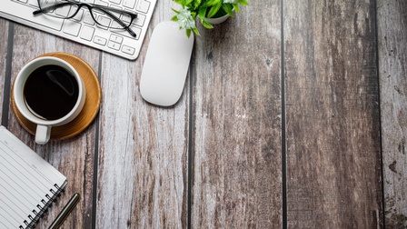 Perspektive von oben auf einen Holzuntergrund mit Kaffee, Tastatur, Maus, Brille sowie Notizbuch und Stift