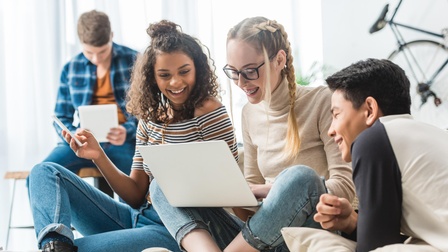 Drei lachende junge Personen auf Couch sitzend und auf Laptop blickend, im Hintergrund weitere Person auf Tisch sitzend