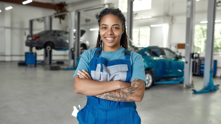 Lächelnde Person mit verschränkten Armen in blauer Arbeitslatzhose steht in Autowerkstatt, im Hintergrund Autos
