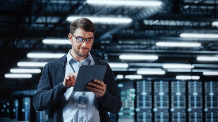 Person mit Brillen blickt auf Tablet, das sie in Händen hält, im Hintergrund verschwommen Serverracks