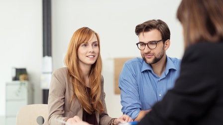 Zwei Personen sitzen an Tisch, eine Person lächelt sanft, die andere trägt Brillen, im Vordergrund verschwommen weitere Person