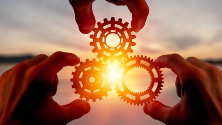 Drei Hände fügen vor einem Sonnenuntergang drei Zahnräder ineinander