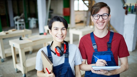 Zwei lächelnde Personen in blauen Arbeitslatzhosen mit Stiften und Notizblöcken in Tischlerwerkstatt stehend