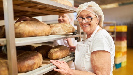 Lächelnde Person mit Brille und Haarnetz sowie in weißem Arbeitsgewand steht neben einem Wagen, auf dem auf mehreren Ebenen verschiedene Brot liegen. Hinter dem Wagen verschwommen steht eine weitere Person.
