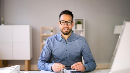 Lächelnde Person mit Brille sitzt an einem Schreibtisch und arbeitet mit aufgeschlagener Mappe, Taschenrechner, im Hintergrund befinden sich Regale und Ablagen