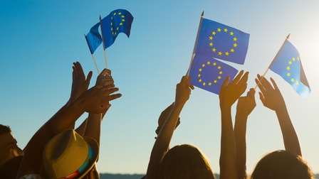 Mehrere Personen  mit hochgestreckten Armen wacheln kleine EU-Flaggen in der Luft, im Hintergrund blauer Himmel