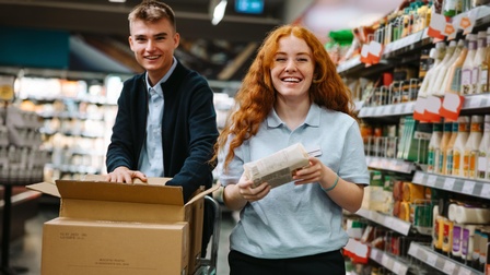 Porträt zweier nebeneinander stehender jugendlicher Personen. Eine Person greift in einen geöffneten Karton, der vor ihr erhöht auf einem Rollwagen steht. Die andere hält eine rechteckige Packung. Rechts daneben ist ein mit Lebensmitteln befülltes Regal.