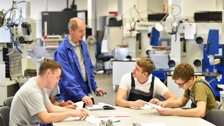 Um einen Tisch sitzen vier junge Personen, die jeweils einen Zettel halten. Zwei von ihnen tragen Latzhosen. Daneben steht eine weitere Person, die etwas erklärt