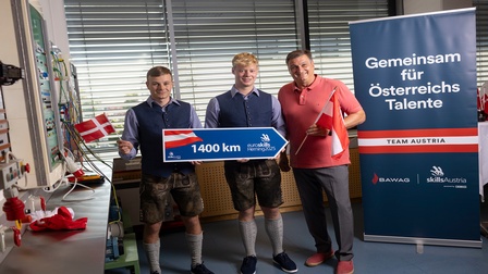 In einem Raum stehen zwei jugendliche und eine erwachsene Person. Die beiden Jugendlichen halten ein Schild, auf dem unter anderem Euroskills steht. Neben ihnen ist ein Banner mit dem Spruch Gemeinsam für Österreichs Talente