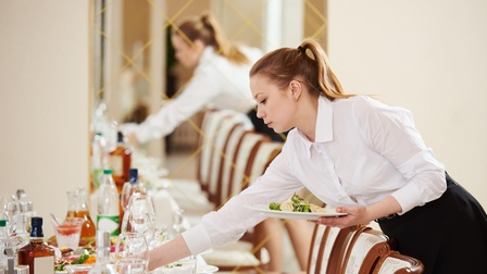 Person in weißer Bluse beugt sich über gedeckten Tisch und greift nach Schüssel mit einer Hand, in anderer Hand hält sie Teller mit Essen