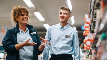 Ein fröhlich Lehrling und seine Ausbilderin beide in hellblauen Arbeitshemden stehen in einem Geschäft neben einem Regal mit Preisschildern und unterhalten sich.