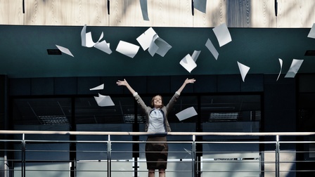 Eine Person im Business-Outfit steht auf einer Galerie in einem modernen Gebäude, wirft zahlreiche Papierblätter jubelnd in die Luft und streckt dabei beide Arme nach oben.