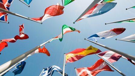 Mehrere Nationalflaggen aus verschiedenen Ländern wehen an hohen Masten im Wind vor einem klaren blauen Himmel