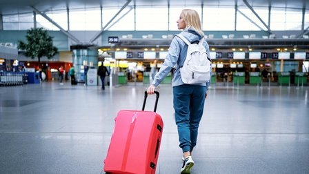 Rückenansicht einer Person, die gerade einen Flughafen betritt und ihren roten Koffer mit sich zieht. Am Rücken trägt sie einen weißen Rucksack und schaut auf die Seite.