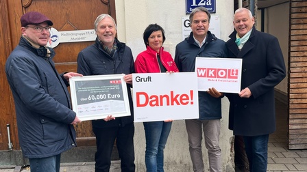 Porträt von fünf Personen, die nebeneinander vor einem Gebäude im Freien stehen. Drei der Personen halten jeweils ein Schild in die Kamera. Auf dem mittleren stehen Gruft und Danke. Auf dem linken steht unter anderem 60.000 Euro