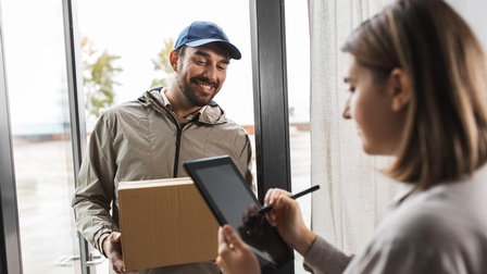 Eine Person mit Jacke und Kappe steht in einer geöffneten Glastür und hält einen Karton. Ihr gegenüber steht eine andere Person, die auf einem Tablet unterschreibt