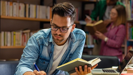 Eine Person sitzt an einem Tisch. In der linken Hand hält sie ein aufgeklapptes Buch. Mit der rechten Hand notiert sie etwas auf einem weißen Blatt. Im Hintergrund sind viele Bücher in Regalen. Daneben steht eine weitere Person