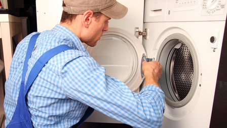 Person in blauer Arbeitskleidung schraubt am Bullauge einer Waschmaschine