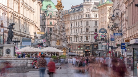 Der Graben in Wien mit seinen historischen Gebäuden sowie zwei Statuen wie der Pestsäule. In der Fußgängerzone gehen und stehen Menschen