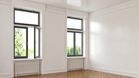 Ein leerer Raum mit Holzboden, weißen Wänden und zwei Doppelfenstern. Unter jedem Fenster ist ein Heizkörper. Hinter den Fenstern sind grüne Bäume