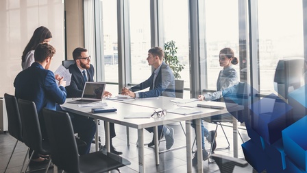 Gruppe an Personen in Businesskleidung befindet sich rund um einen Besprechungstisch in einem modernen und hellen Besprechungsraum. Rechts im Bild sind blaue Würfel darüber gelegt