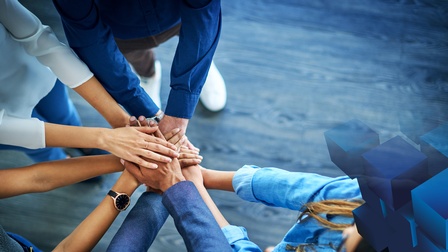 Fünf Personen stehen in einem Halbkreis. Jede Person hält beide Hände in die Mitte des Halbkreises. Die Hände liegen übereinander. Rechts im Bild sind blaue Würfel darüber gelegt