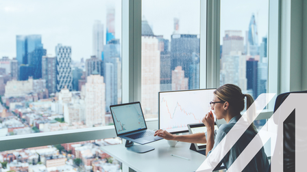 Rückansicht einer Business-Frau vor einer Glasfront eines Büros in einem Wolkenkratzer mit Blick auf eine moderne Großstadt. Vor ihr ein Computer mit finanziellen Graphen und Statistiken auf dem Monitor.