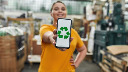 Nahaufnahme von einem Smartphone mit grünem Recyling Icon, das von einer Person mit orangem T-Shirt gehalten wird