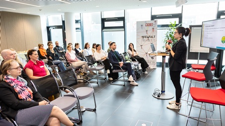 Person in Seitenansicht steht auf einer Bühne und spricht in ein Mikrofon einem sitzenden Publikum zugewandt, im Hintergrund Rollup mit Schriftzug Vielfalt vernetzt