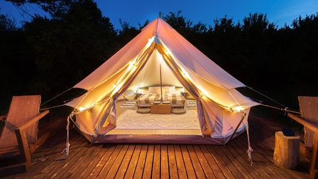 Auf verlegten Holzbrettern steht ein großes Zelt, in dem ein Teppichboden, ein Bett und Lichter sind. Hinter dem Zelt sind ein Wald und der dunkle Himmel