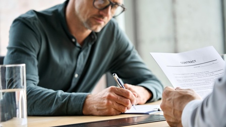Person mit Brillen unterfertigt Dokument, Ausschnitt anderer Person, die Dokument mit Schriftzug Contract in Händen hält 