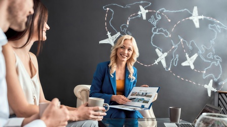 In der linken Bildhälfte sitzen zwei Personen an einem Tisch. Neben ihnen sitzt eine weitere Person, die einen aufgeklappten Ordner hält, auf dem Best Places steht. Hinter dieser Person ist eine dunkle Wand, auf der die Kontinente der Welt sind