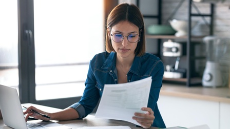 Person mit Brillen sitzt an Tisch und blickt auf Dokument, nebenstehend aufgeklapptes Notebook, auf das Person eine Hand mit Stift auf Tastatur hält
