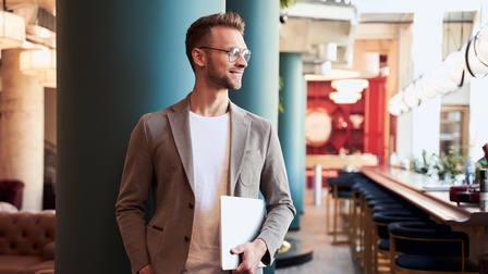 Lächelnde Person mit Brillen hält Notebook in der Hand und steht vor blauer Säule in einem Lokal, im Hintergrund Bar mit Hockern