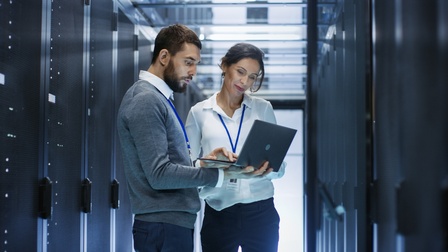 Zwei Personen stehen vor Servern und blicken auf aufgeklapptes Notebook, das eine Person in Händen hält