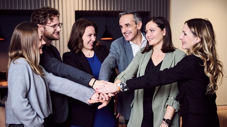 Eine Gruppe von Personen steht zusammen. Jede hat eine Hand ausgestreckt, die Personen führen die Hände in der Mitte zusammen und stapeln diese übereinander. 