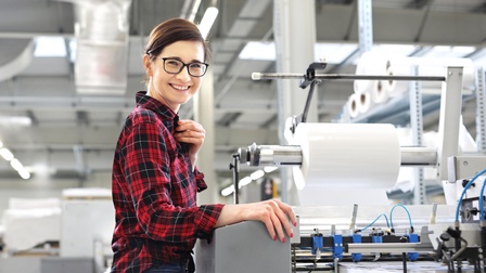 Eine Person mit Brille steht seitlich vor einer großen Maschine. Auf einer Fläche der Maschine liegt ein hoher, weißer Papierstapel. Schräg über dem Stapel ist eine Metallrolle, auf der eine dicke, weiße Rolle ist. Die Person blickt direkt in die Kamera.