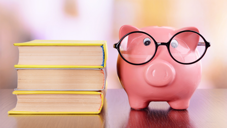 rosa Sparschwein mit schwarzer runder Brille steht neben Bücherstapel auf einem Tisch