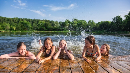 Fünf Kinder planschen und lachen an einem Badesee an einem sonnigen Sommertag.