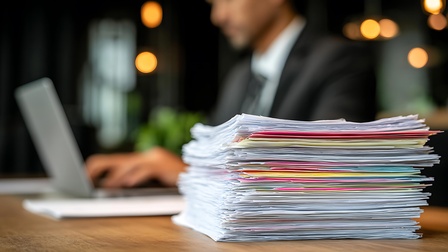 Papierstapel im Vordergrund, Mann am Laptop im Hintergrund