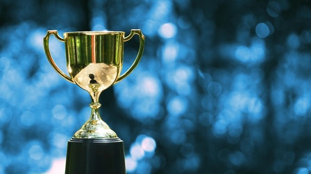 Goldener Pokal steht auf einem Holztisch, im Hintergrund zeigt sich ein blaues Bokeh, Konzept für Sieg